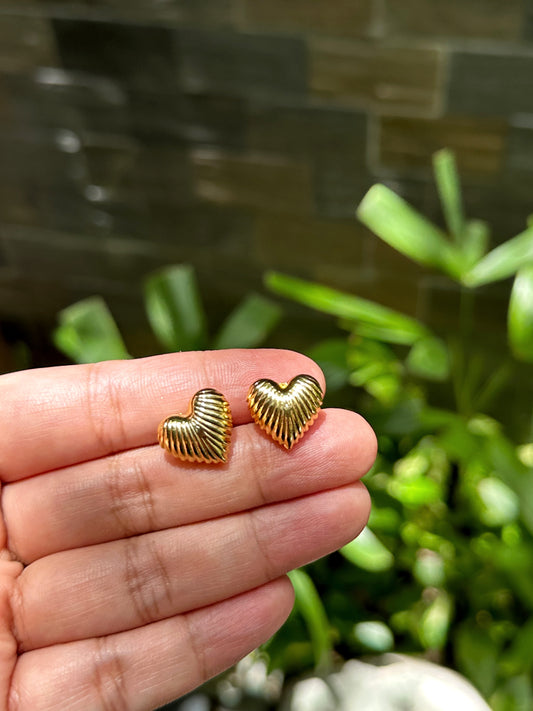 Hand holding a pair of gold-tone ribbed heart stud earrings with garden background