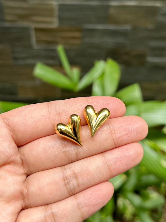 Gold puff heart earrings held by hand in front of greenery background