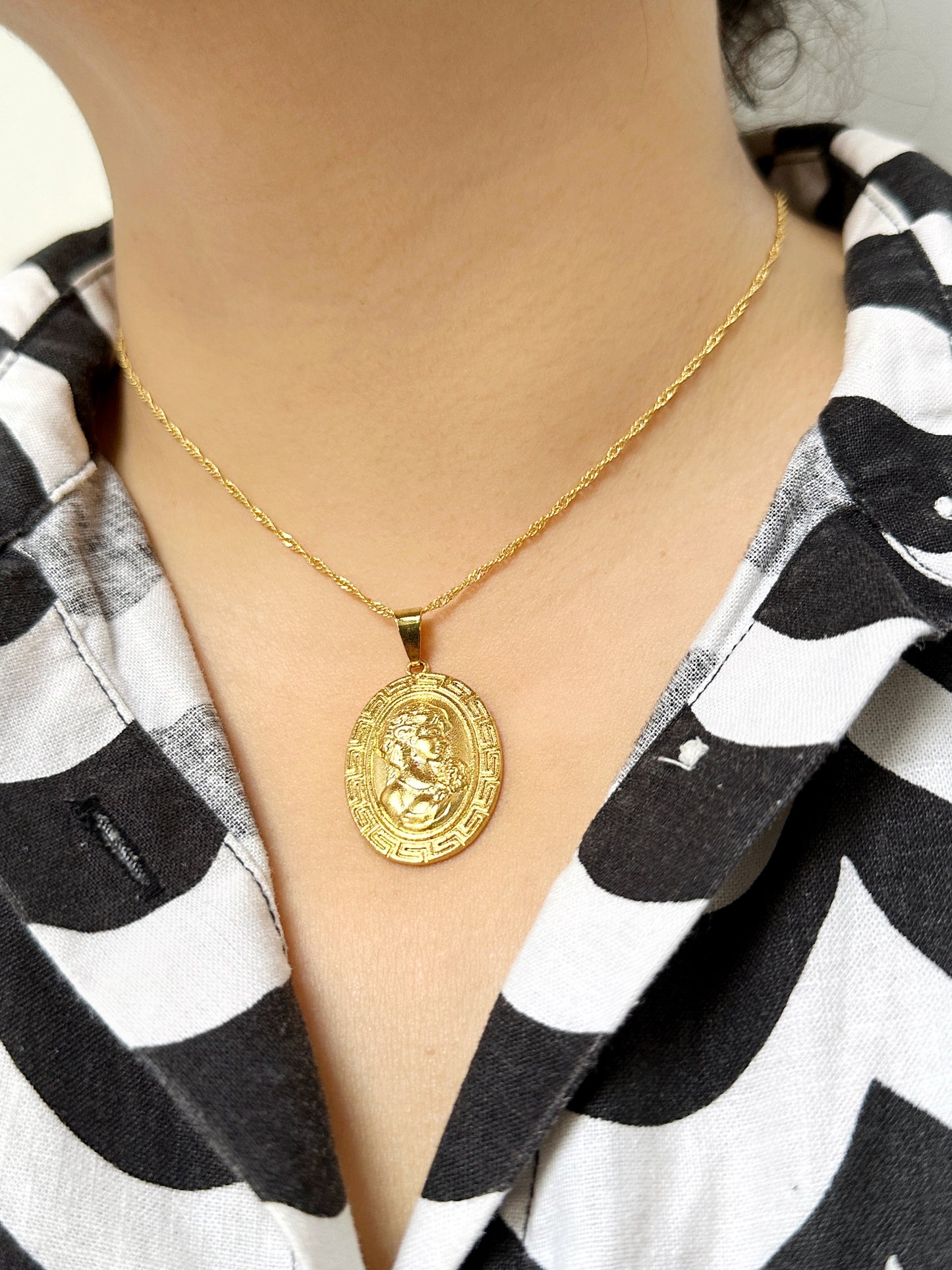 Close-up of a woman wearing a gold medallion pendant necklace on a rope chain, featuring a Greco-Roman profile design