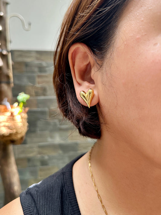 Close-up of a woman wearing a gold-tone flirty heart stud earring made of stainless steel, styled with a short haircut and a dainty chain necklace in an outdoor setting.