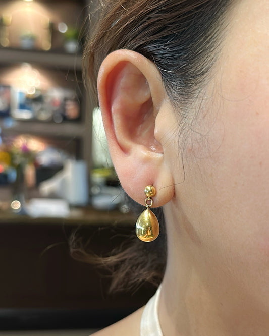Gold teardrop earring worn by a person with a blurred indoor background
