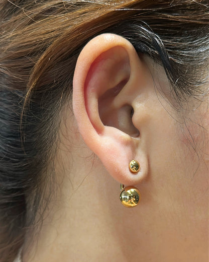 Close-up of an ear wearing gold round sphere front-back earrings with hair partially covering the frame.